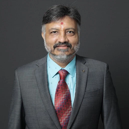 Head-and-shoulders board portrait of Sanjay Ajmera in a grey suit and patterned tie against a dark studio background.